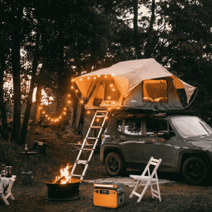 Estación de energía portátil LiFePO4 naranja en un campamento con fogata y coche con tienda de campaña de techo.