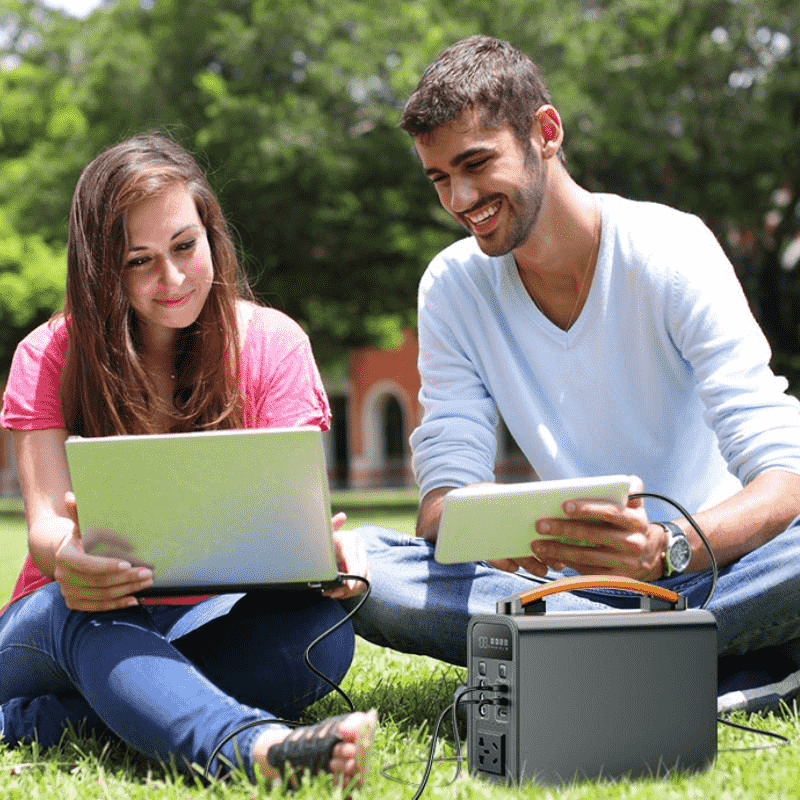 Pequeña estación de energía portátil alimentando una computadora portátil y una tableta en el césped durante actividades al aire libre.
