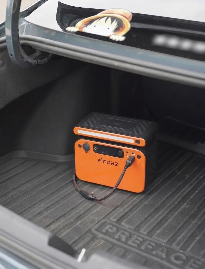 車のトランクの床面に置かれ、ケーブルで充電されているPiforzなどのオレンジ・黒のポータブル電源。