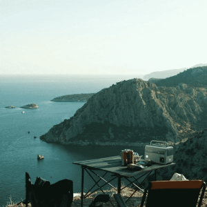 A compact portable power station with AC outlets on a camping table overlooking a scenic sea and mountain landscape at dusk.