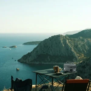A compact portable power station with AC outlets on a camping table overlooking a scenic sea and mountain landscape at dusk.