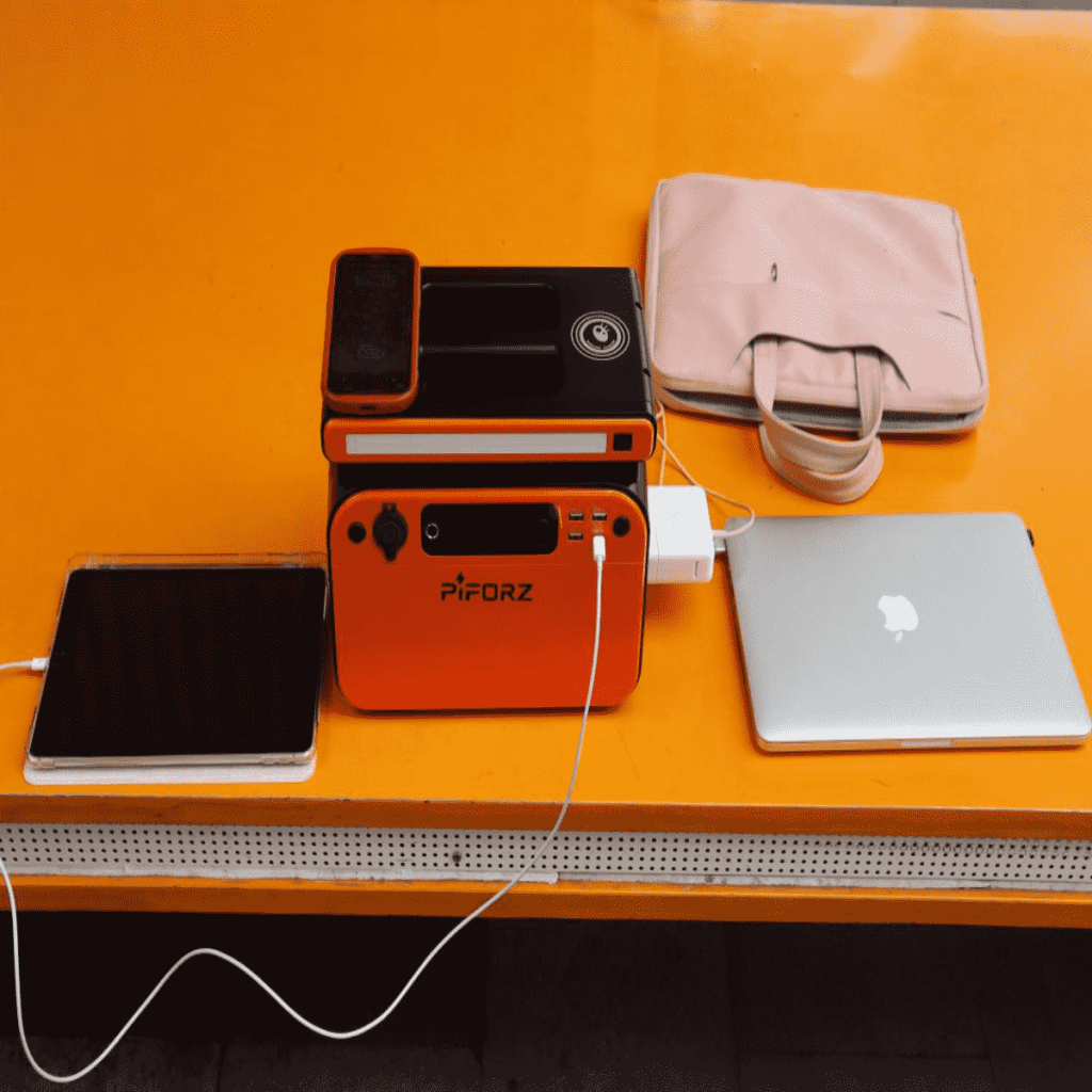 A compact orange PiForz solar powered emergency generator on a table, simultaneously charging a laptop, tablet, and smartphone.