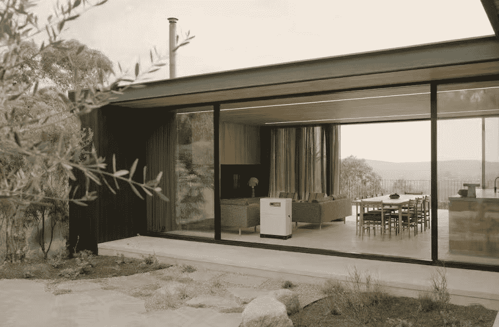 A standalone whole-home energy storage battery system placed outside the glass door of a modern residential house with limited roof solar suitability