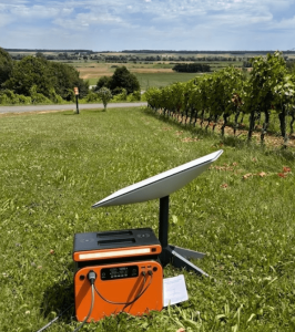 An orange portable power station connected via a cable to power a Starlink Mini satellite dish set up on a grassy lawn next to a vineyard.