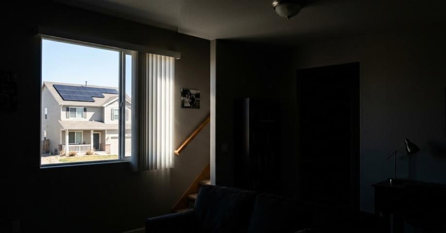 View from inside a dark room looking out at a neighbor's roof covered in solar panels on a bright sunny day during a grid outage.